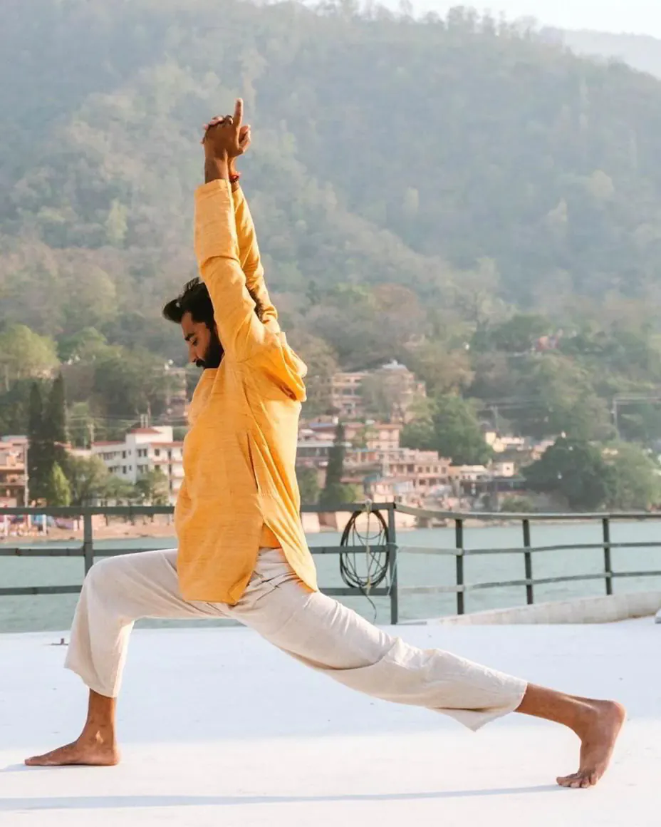 A man in a golden kurta in a high lunge with arms overhead on a rooftop above the Ganga