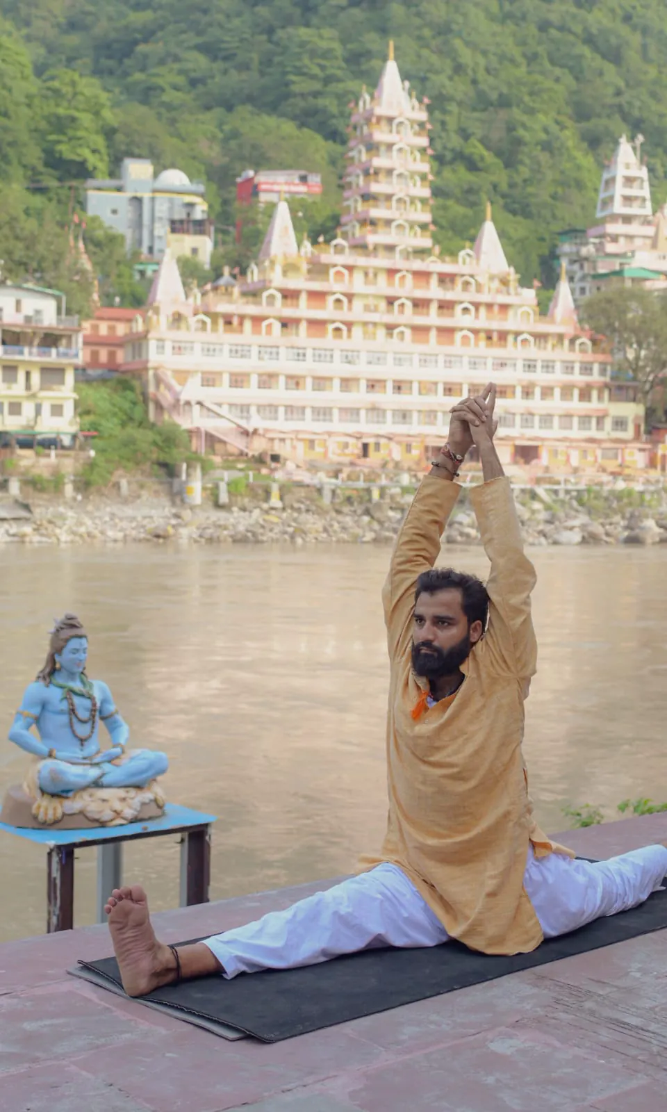A man in a seated wide-leg split with palms raised on a ghat, with a tiered temple across the Ganga