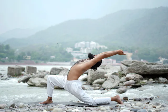 A man in white trousers in a deep back-bend on the rocks of the Ganga, misty green hills behind