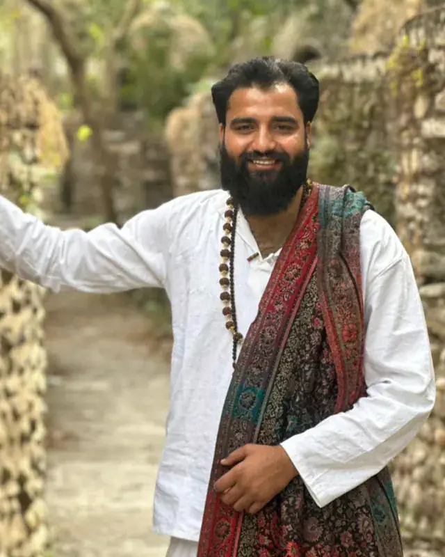 Yogi Arya smiling in a white kurta and patterned shawl, leaning on a stone wall in a lane