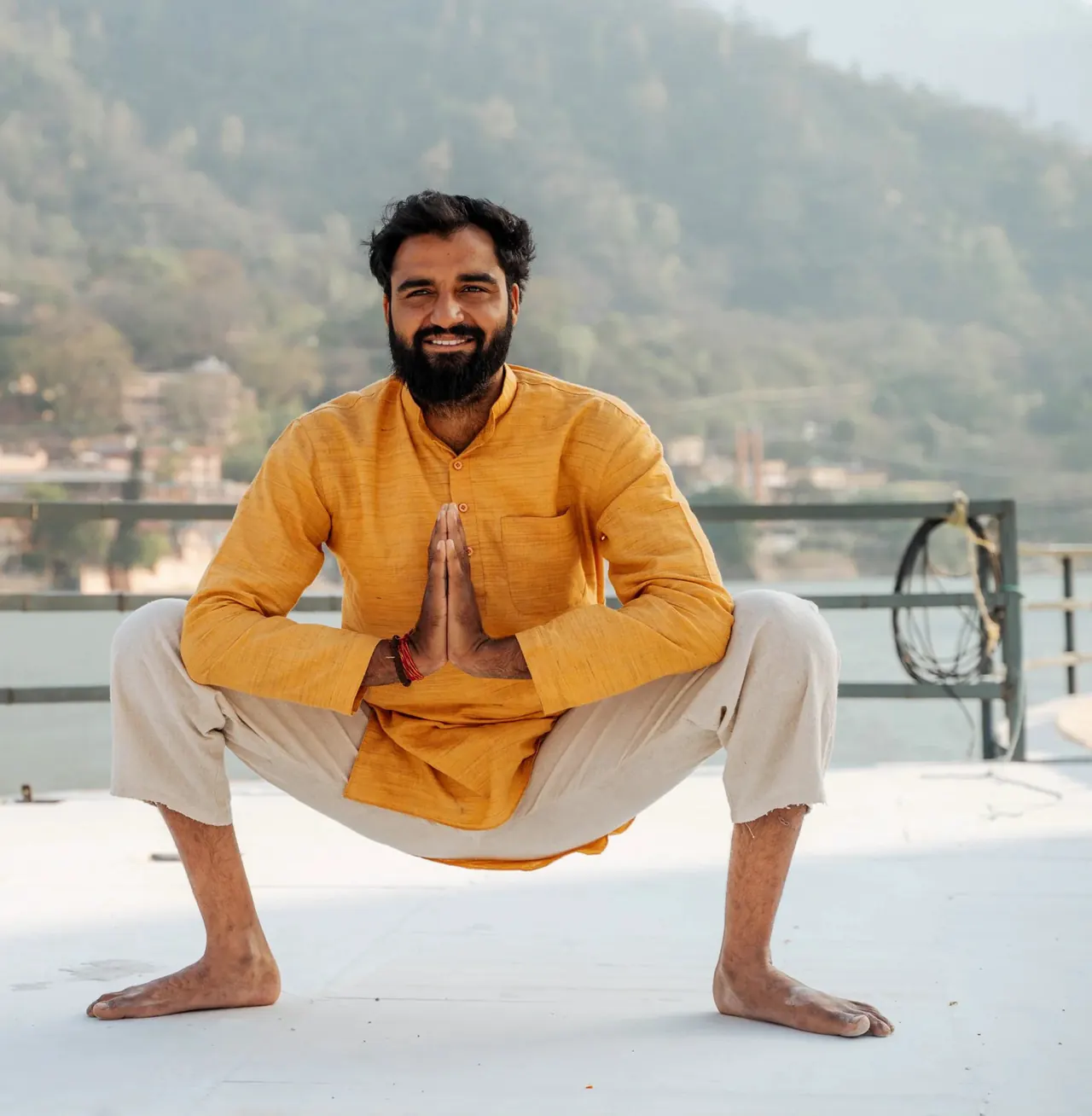Yogi Arya in a deep squat with palms together on a rooftop above the Ganga