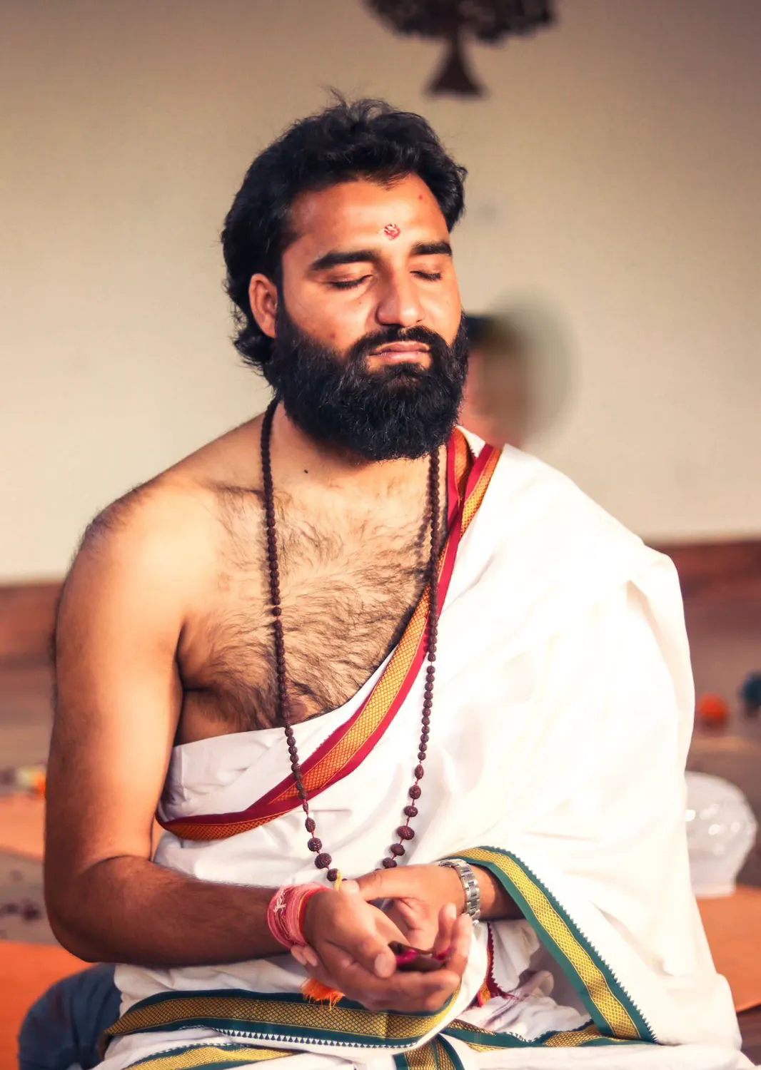 Yogi Arya with eyes closed in prayer, wearing a mala and a white cloth with a red and gold border