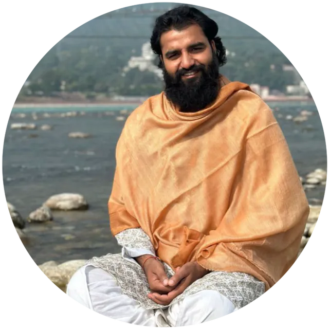 Yogi Arya sitting by the Ganga on river rocks in a gold shawl, smiling