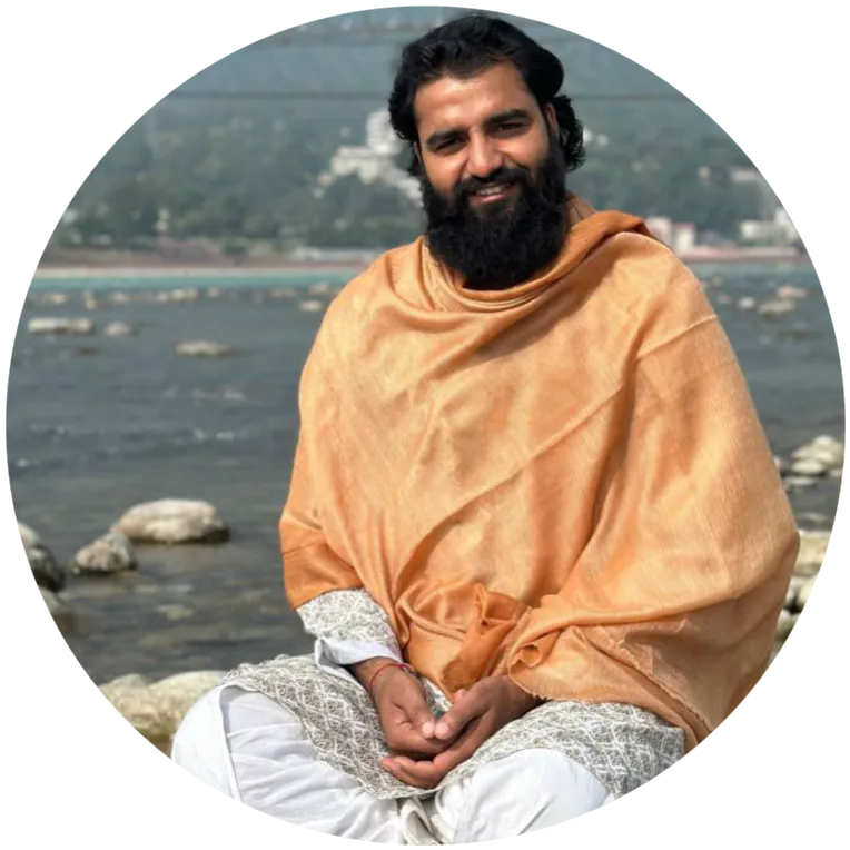 Yogi Arya sitting by the Ganga on river rocks in a gold shawl, smiling