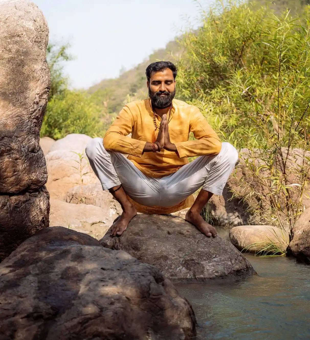 Yogi Arya with eyes closed and palms together, squatting on a river rock among bamboo and boulders