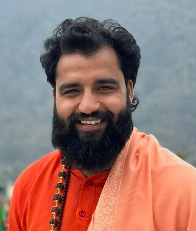 Yogi Arya smiling in an orange kurta and peach shawl, misty hills behind