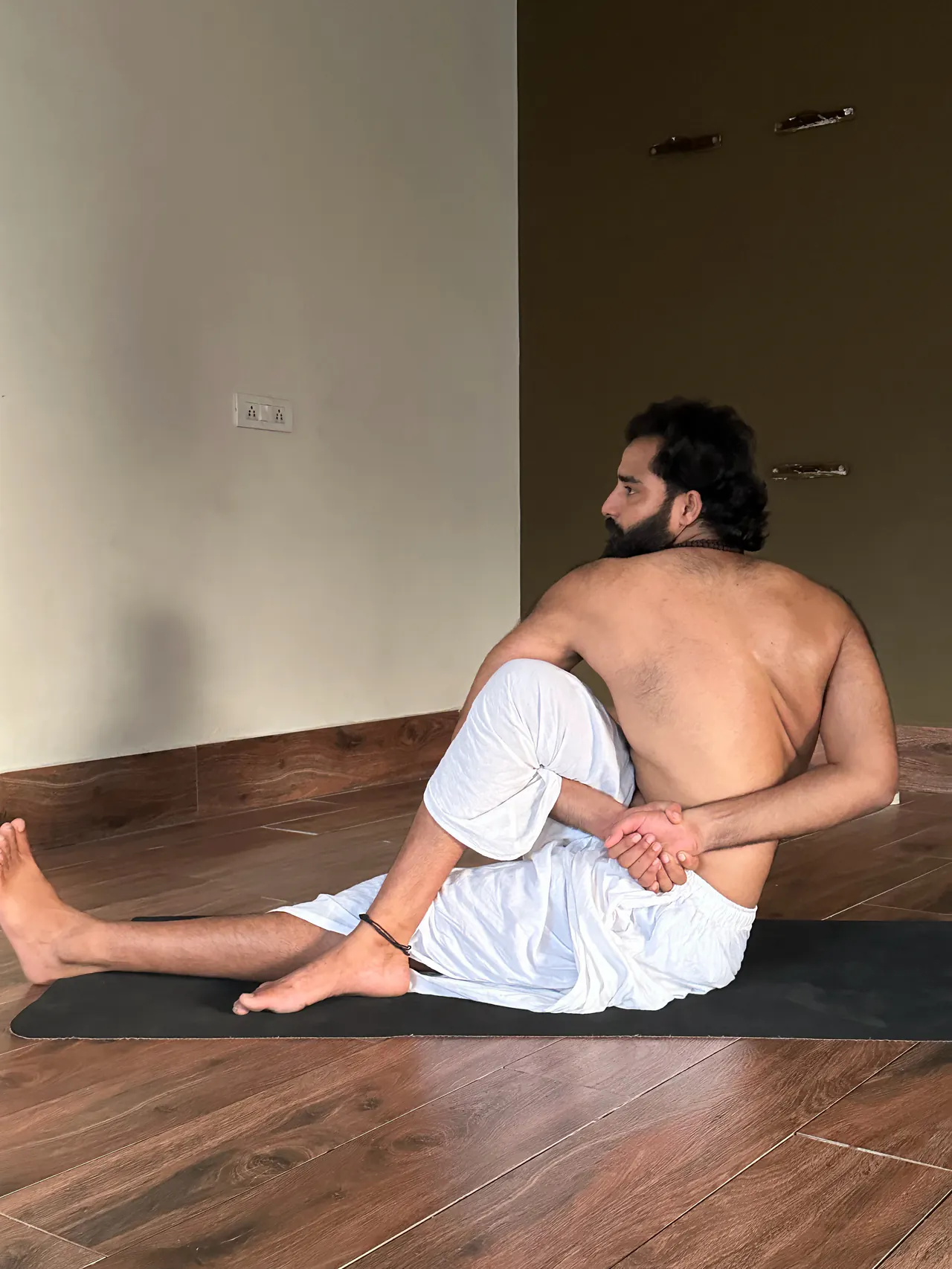 Yogi Arya in a seated twist on a black mat in a quiet hall, seen from the side