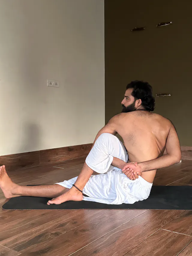 Yogi Arya in a seated twist on a black mat in a quiet hall, seen from the side